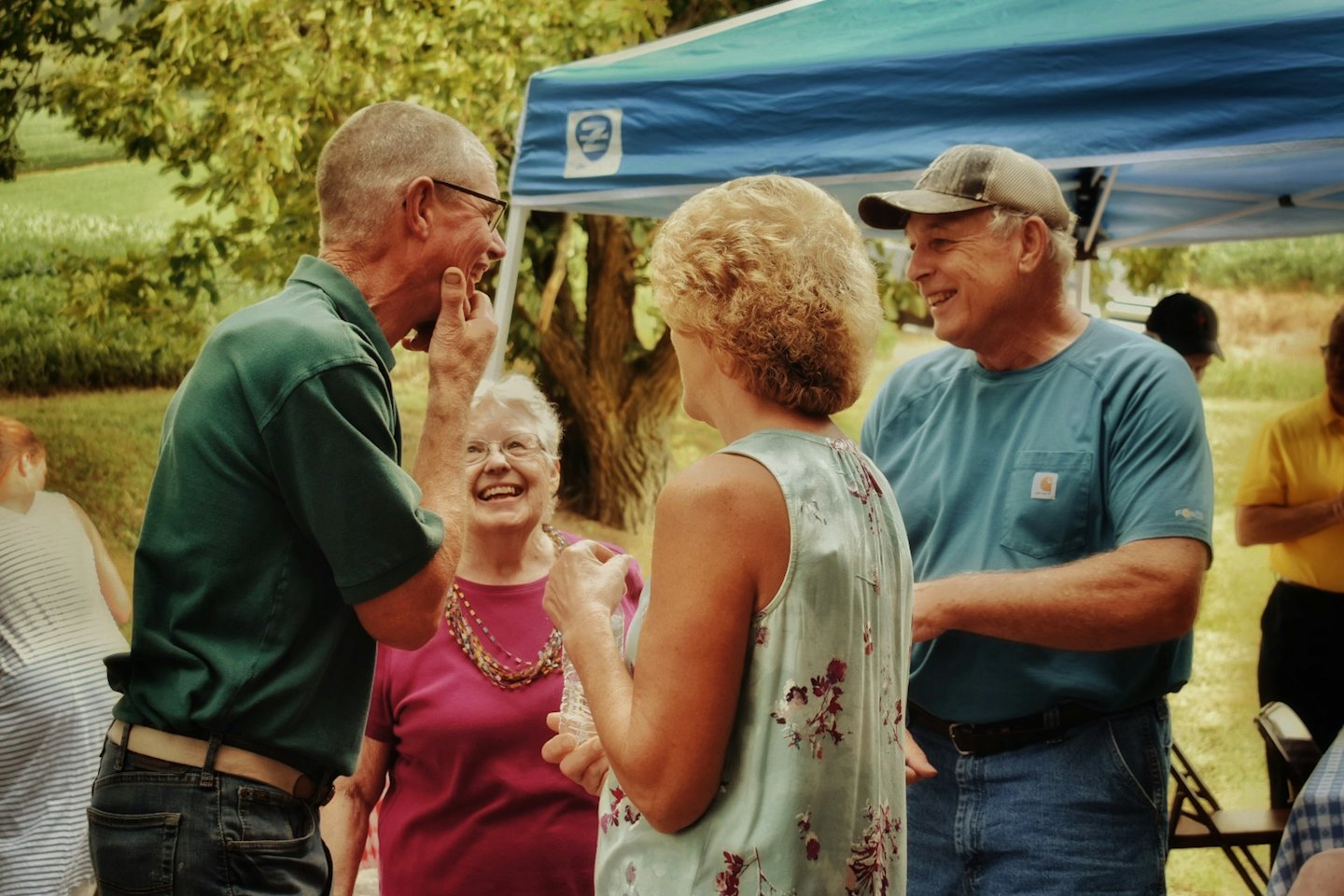 Adults laughing and conversing outdoors under a blue canopy tent.