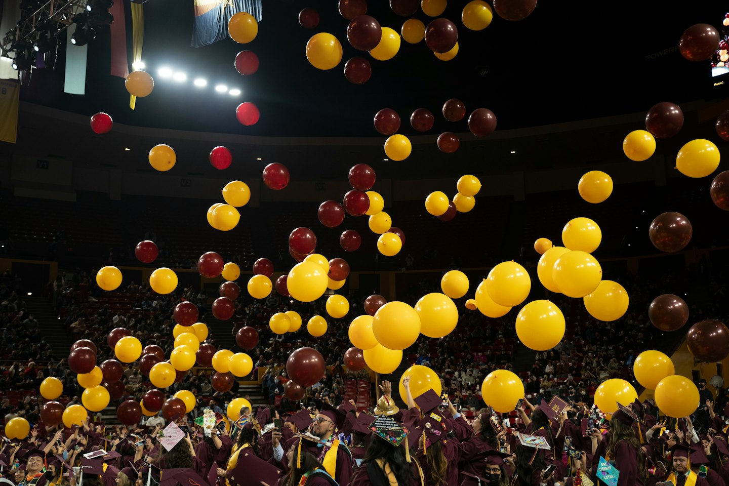 Graduates in maroon gowns look up as balloons drift upward.