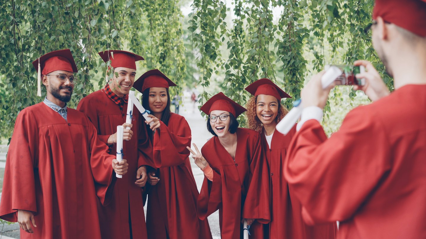 Six graduates in red caps and gowns celebrate outdoors.