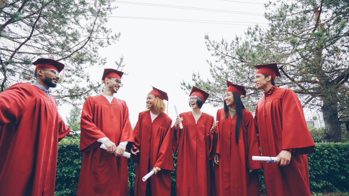 Graduating students in red gowns holding diplomas and celebrating outdoors.